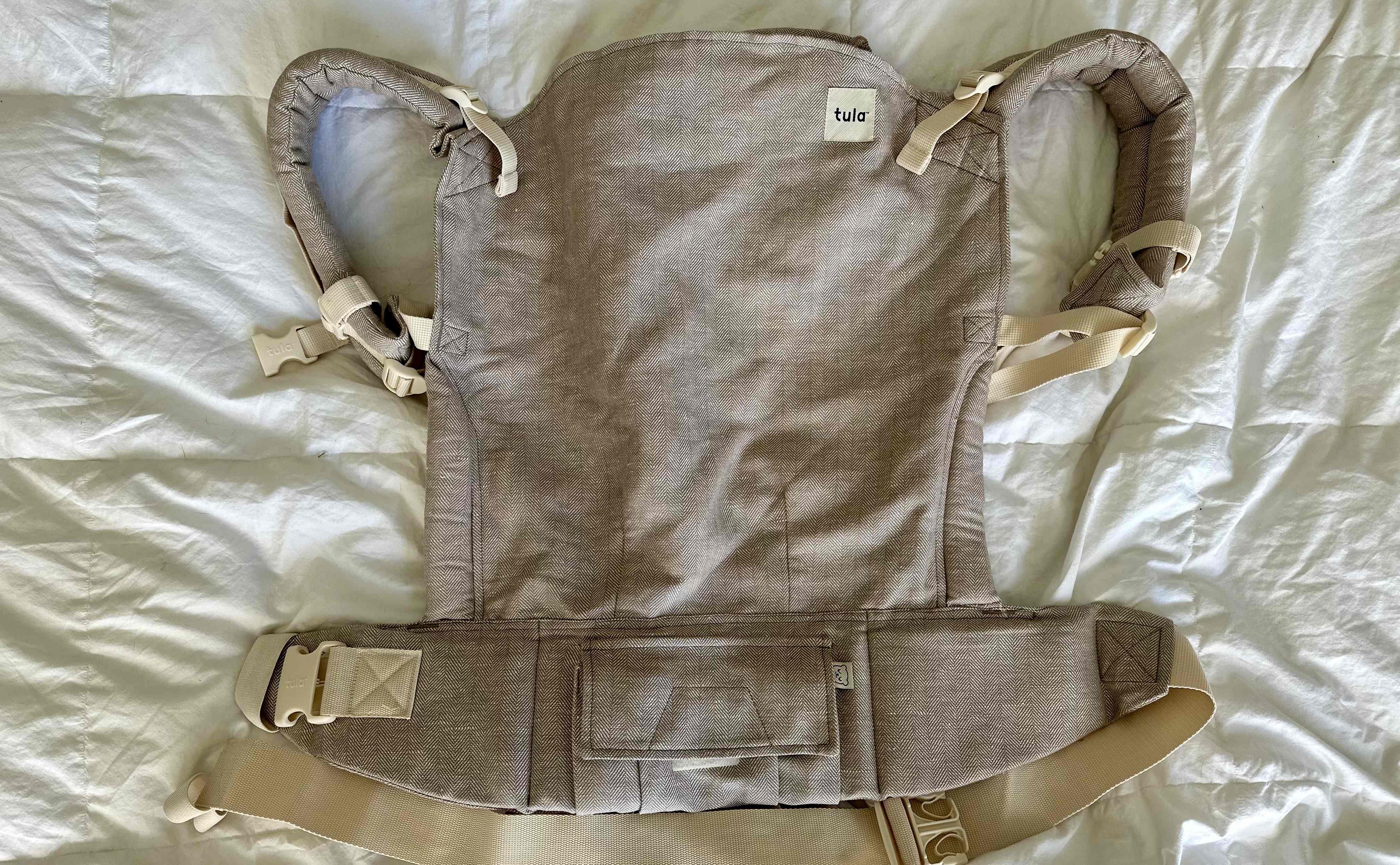 A beige linen Tula Toddler baby carrier laying flat on a white comforter.