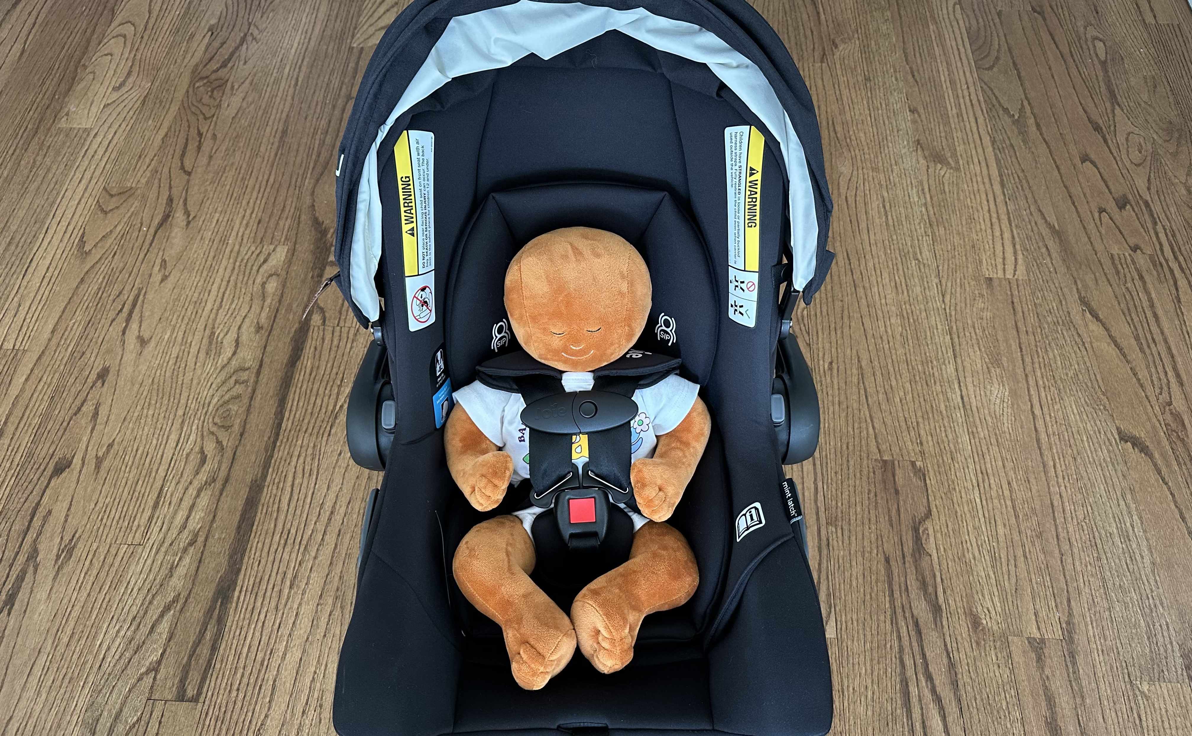 Front view of a small doll harnessed into a black Joie Mint Latch Infant Car Seat.