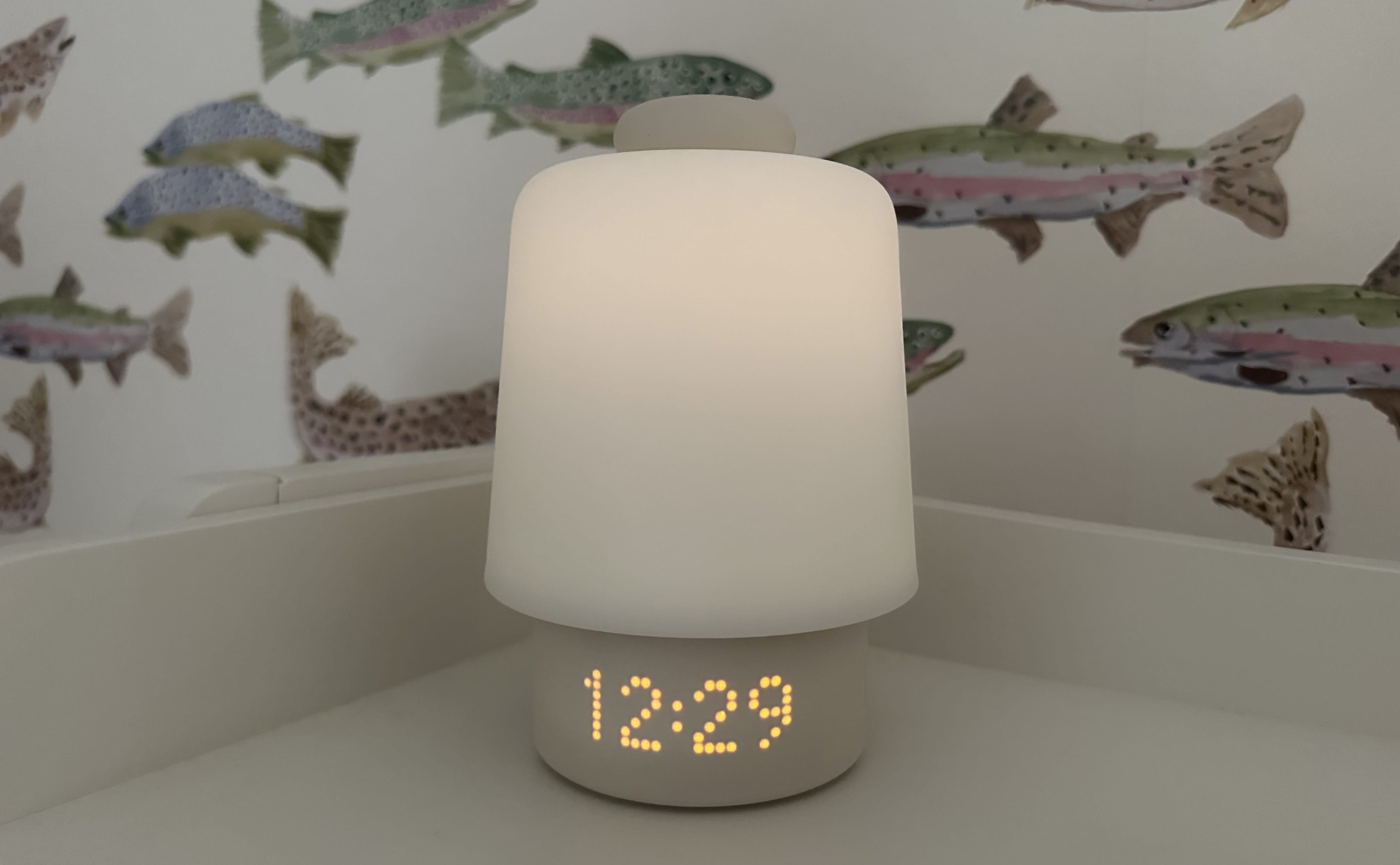 A Hatch Baby sound machine in white, with the night light on, on a white shelf.