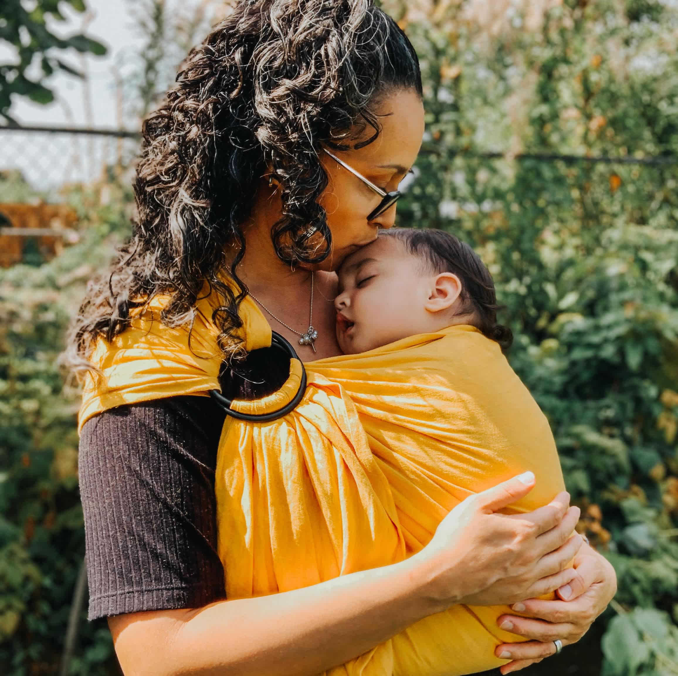 Parent babywearing in a ring sling carrier, kissing their baby's head.