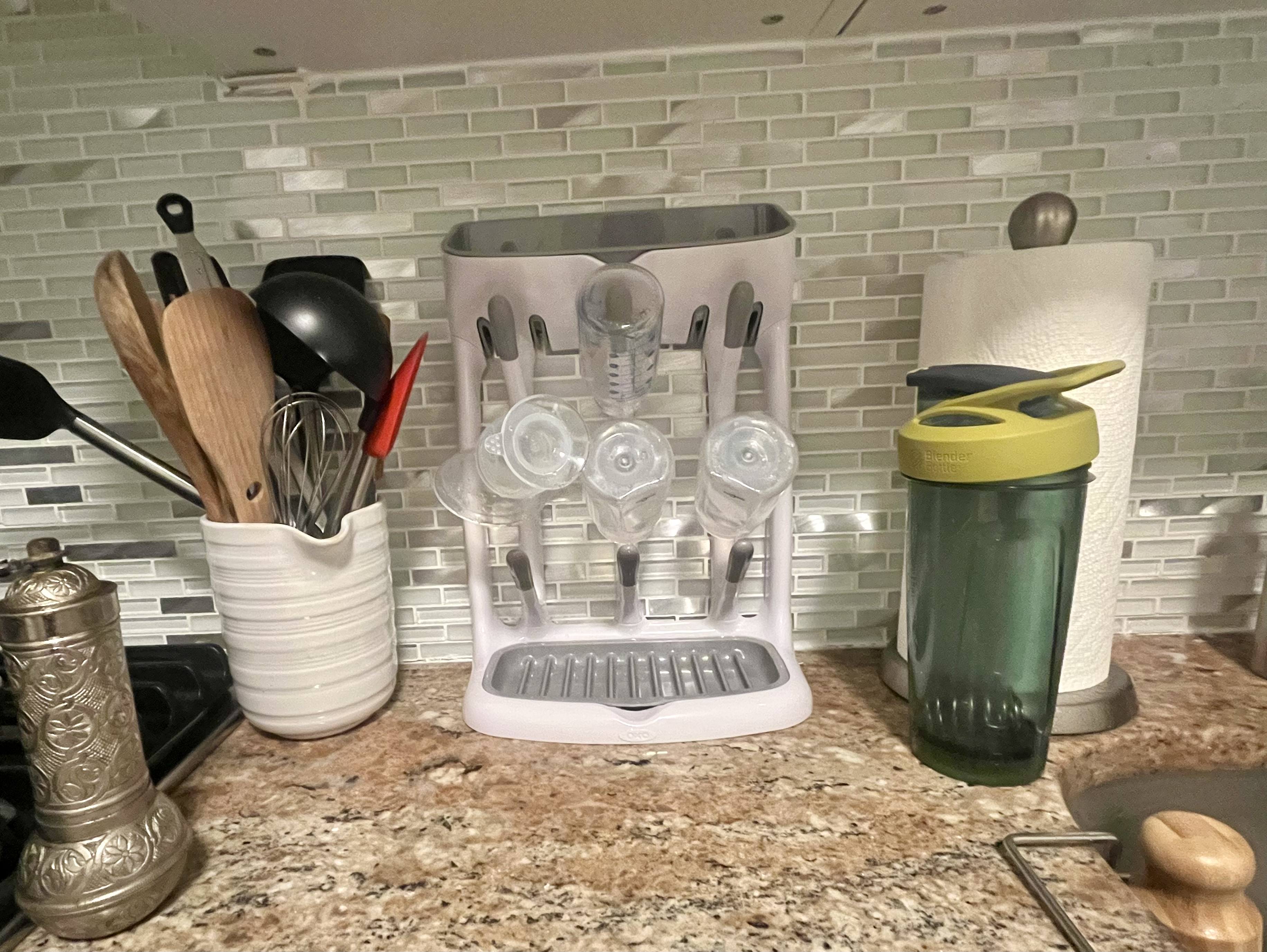Grey and white OXO Space Saving Bottle Drying Rack loaded with several bottles and accessories, on a kitchen counter amongst other items like paper towels and a container of kitchen utensils.
