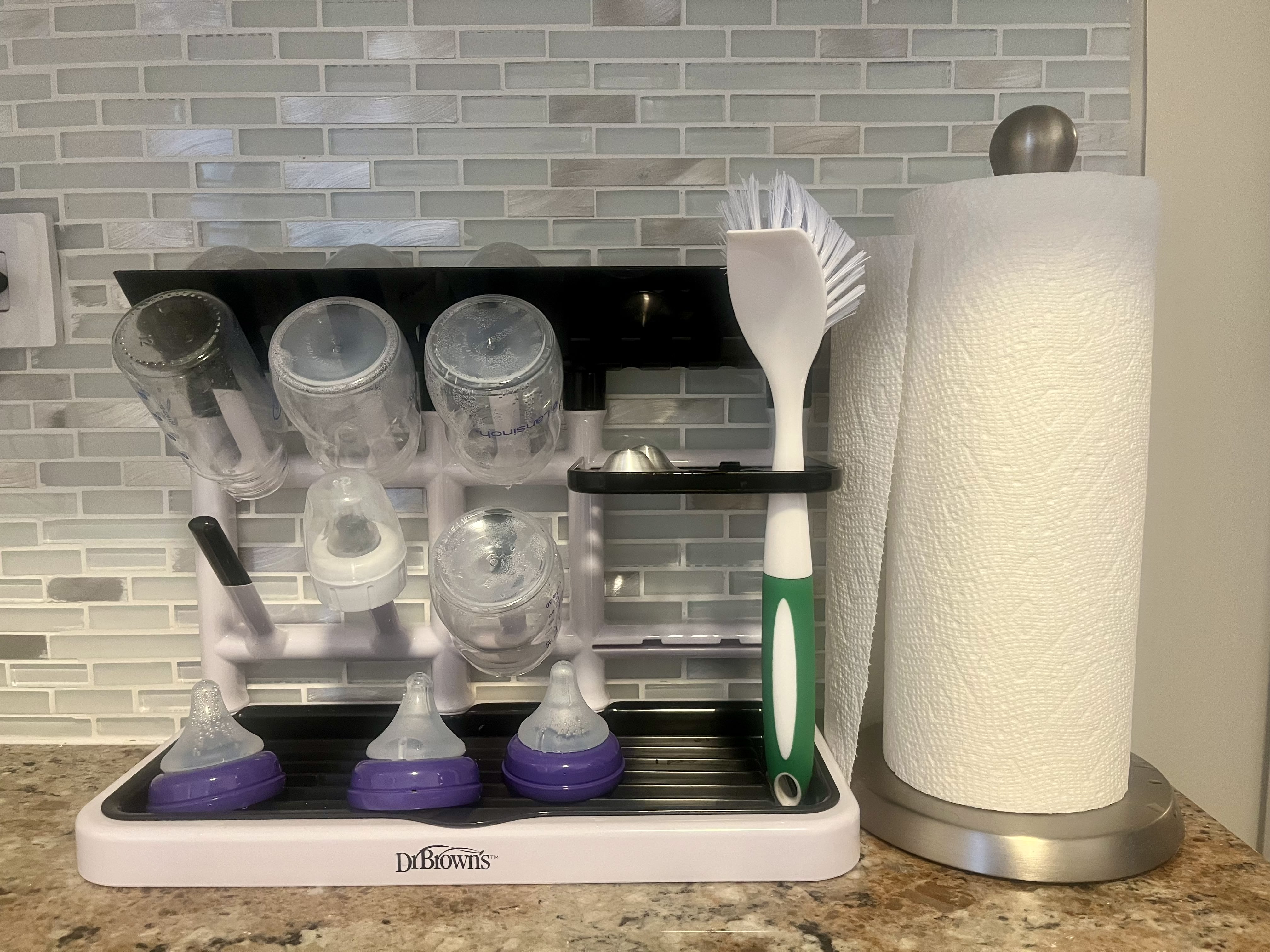 Black and white Dr. Brown's Drying Tower loaded with several bottles and nipples on top of a kitchen counter next to a roll of paper towels.