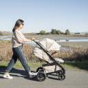 A woman with black hair and sunglasses pushes a Nuna Tavo Next stroller with a beige canopy down a paved path with grass and water in the background. thumbnail.