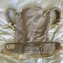 A beige linen Tula Toddler baby carrier laying flat on a white comforter. thumbnail.