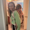 A white woman with blonde hair wearing a young girl on her back in a beige Tula Toddler baby carrier. thumbnail.